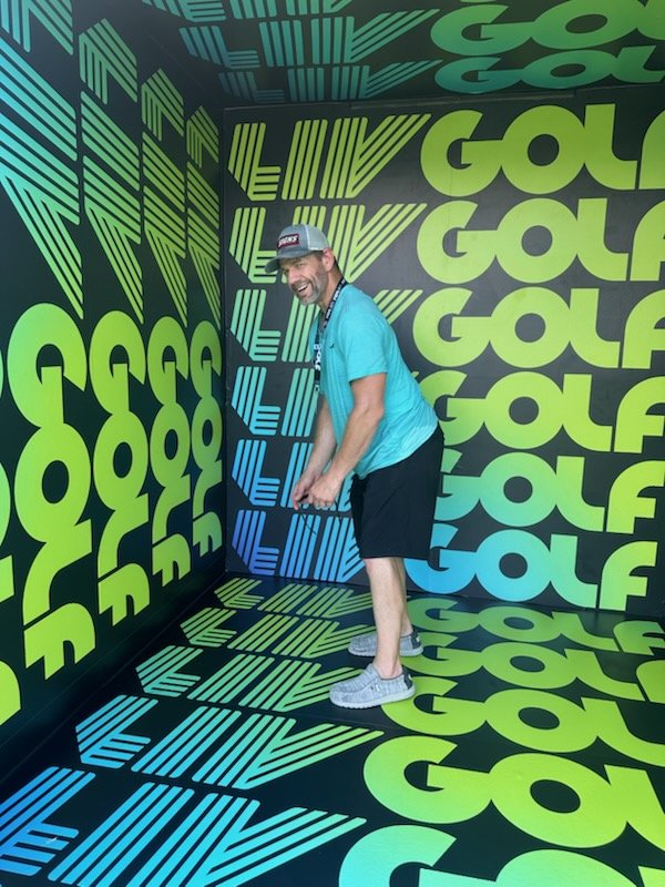 A man smiling and preparing to hit a golf ball in a colorful room with bright green, blue, and black walls and floor covered with the word 'GOLF' in different orientations.