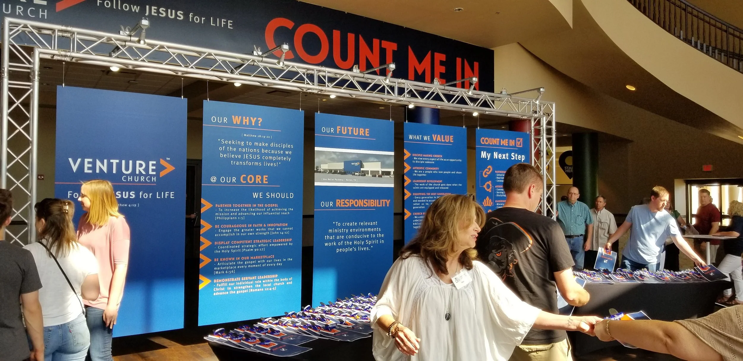 People gathered around a display booth at Venture Church with informational banners about their mission and values, and items spread out on a table.