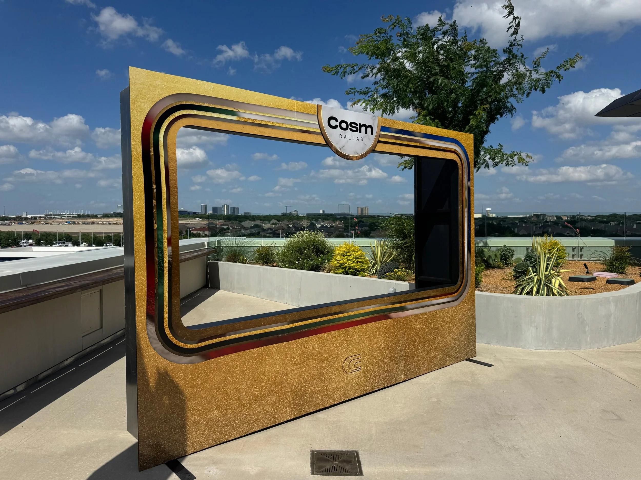 A decorative mirror with a retro design, featuring a gold border and colorful stripes, located on a rooftop with a cityscape in the background under a partly cloudy sky.
