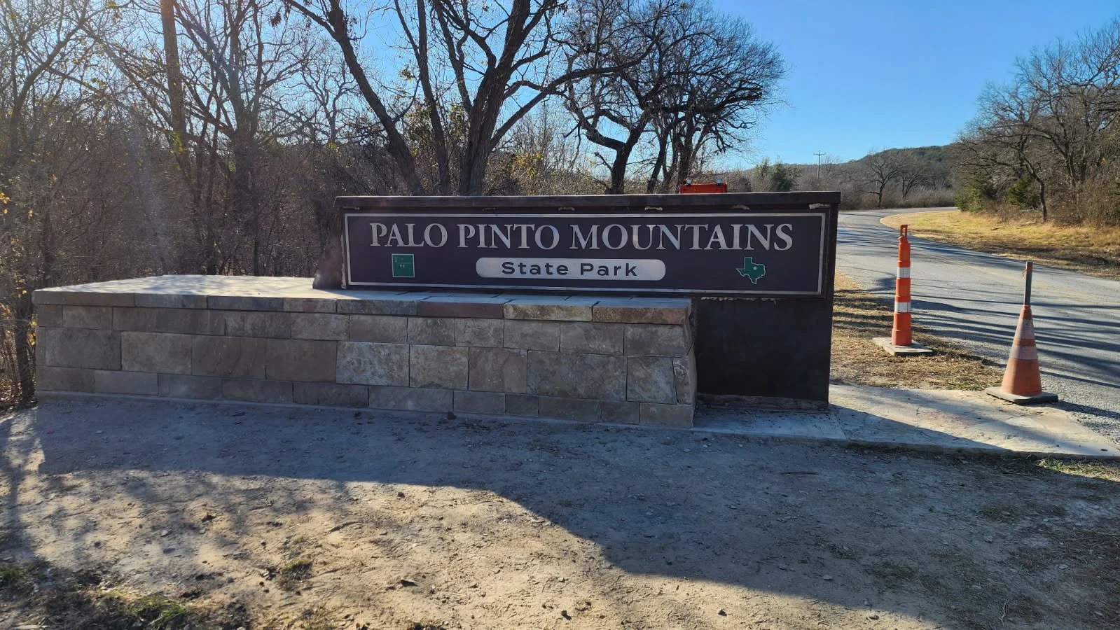 Sign at Palo Pinto Mountains State Park entrance with a stone base, surrounded by trees and a dirt road with traffic cones on the side.