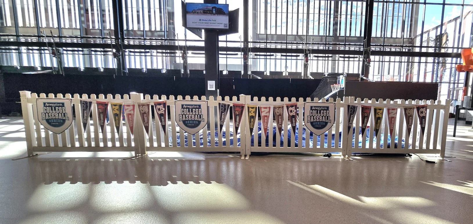 Indoor sports area with a white picket fence displaying Amegy Bank College Baseball Series banners, a basketball hoop, and a scoreboard on a pole.