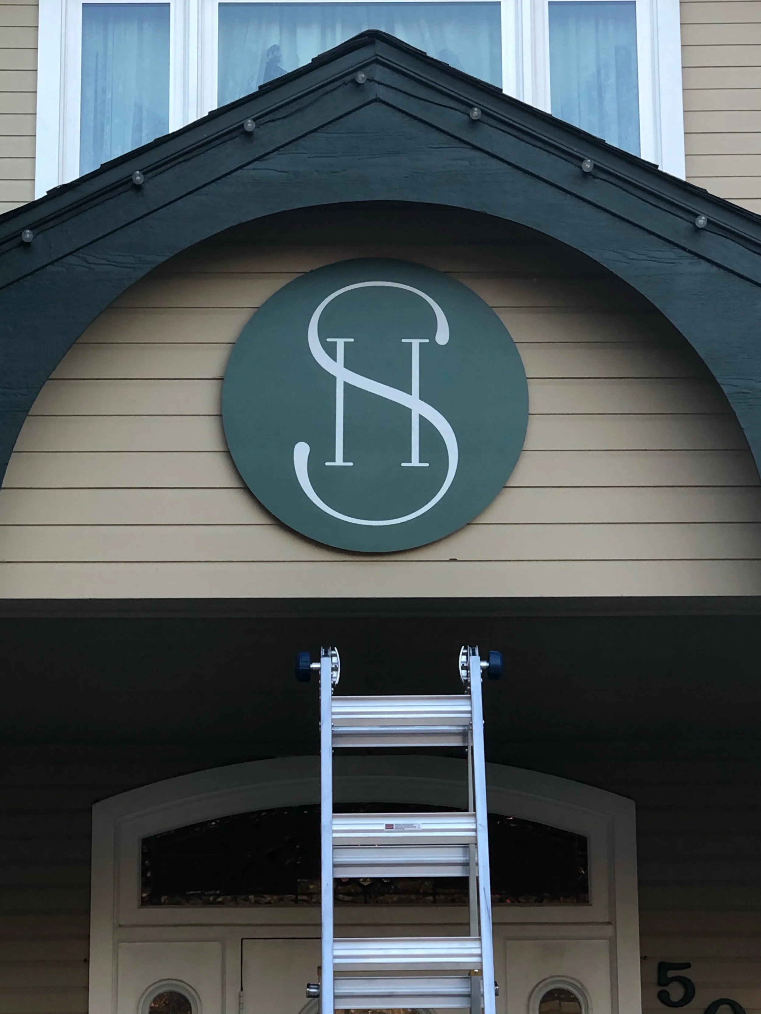 The exterior of a house with a dark green arched awning over a porch and a circular sign with stylized letters 'S' and 'L'. A ladder is set up beneath the sign, extending to the edge of the awning. The house has beige siding and a second-story window with curtains.