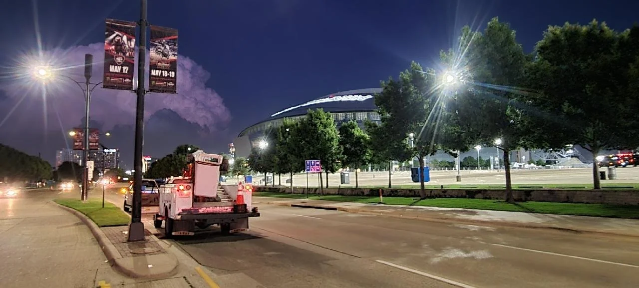 Nighttime view of a street with a lit-up stadium in the background, trees lining the sidewalk, and a maintenance vehicle parked on the road.
