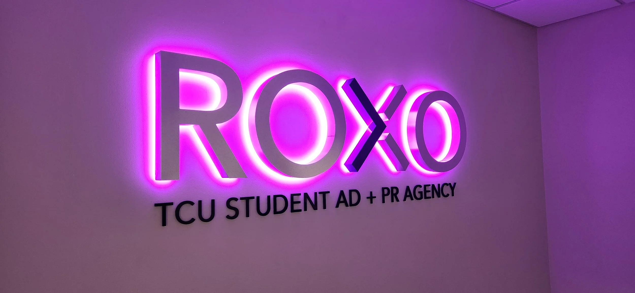 Neon sign that reads 'ROXO' in purple and black letters, with smaller black text underneath that says 'TCU STUDENT AD + PR AGENCY'.