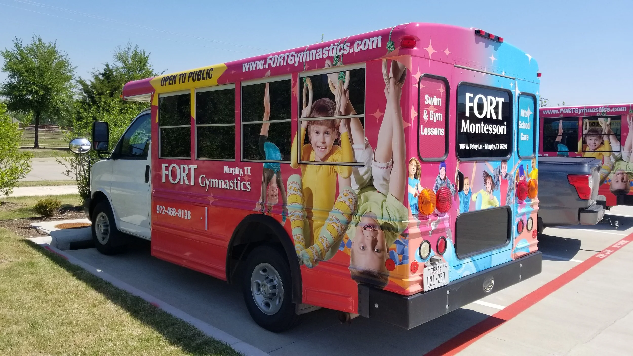 Colorful school bus with graphics of children playing, advertising Fort Gymnastics in Murphy, Texas, parked in a lot with trees in the background.