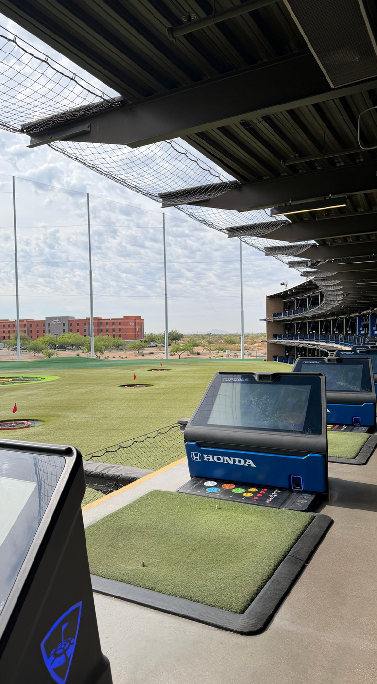 Golf driving range with automated tee stations, including Honda and Topgolf branded screens, under a covered area, overlooking an open field with flags and practice holes.