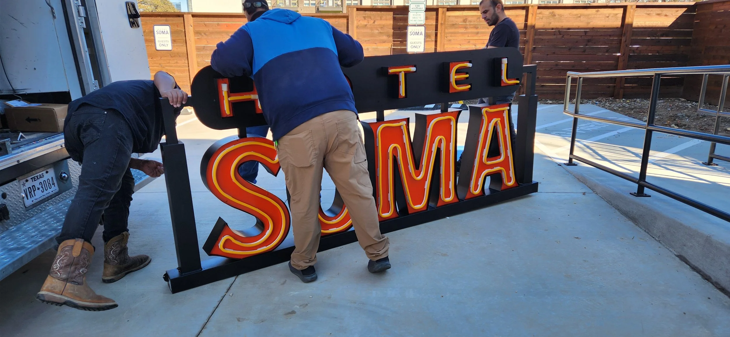 Three people installing a neon sign that reads 'HOTEL SOMA' outside a building. One person is bent over helping, another stands behind, and the third person is observing, with a wooden fence and a parking area in the background.