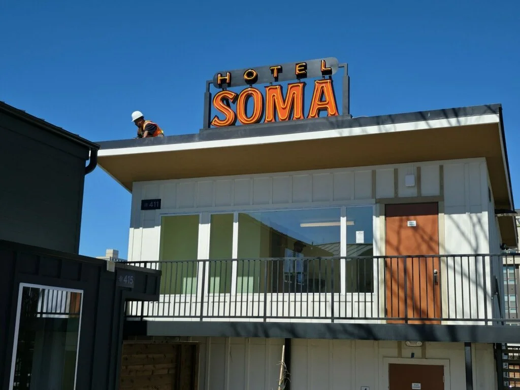 Exterior of a building with a neon sign that reads 'HOTEL SOMA' on the roof, and a construction worker wearing a white helmet working on the roof. The building has a large window, a balcony, and wooden doors.