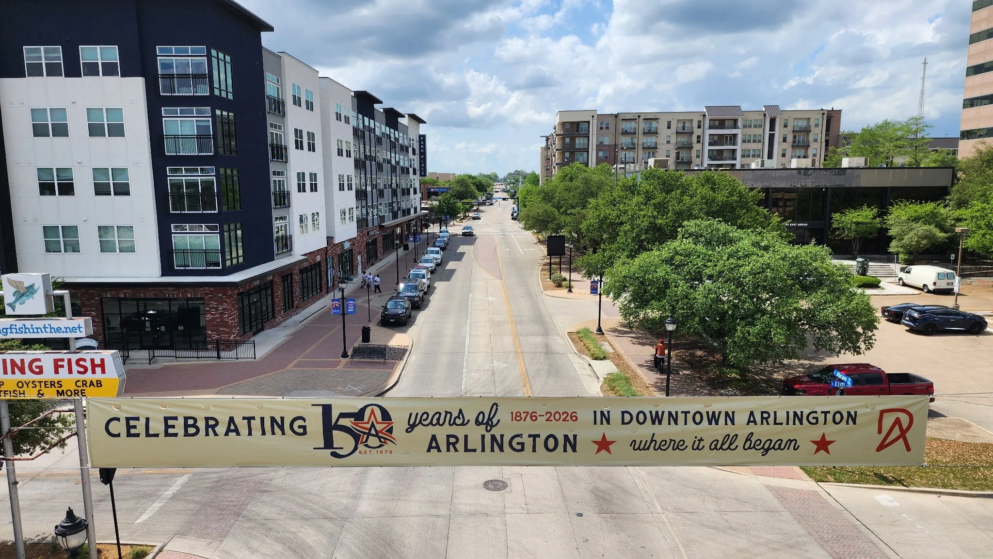 Street view of downtown Arlington with modern apartment buildings, parked cars, and a large banner celebrating 150 years of Arlington.