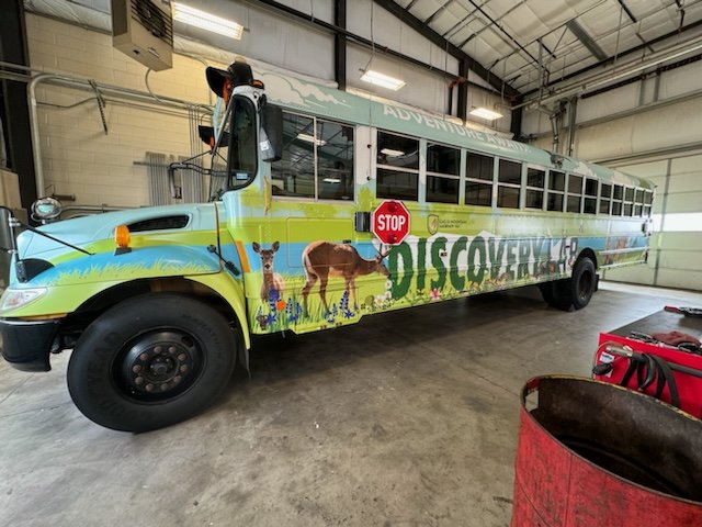 School bus decorated with colorful outdoor adventure mural featuring animals like giraffes and bears, with the word 'DISCOVERY' on the side and a stop sign attached to the front.