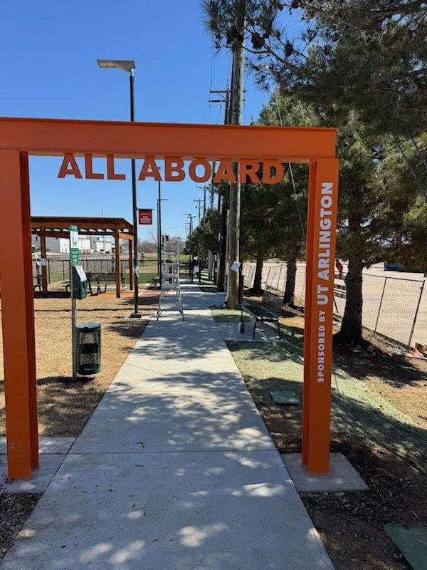 Orange archway with 'ALL ABOARD' in large letters, at a bus stop with benches, trash can, and bus shelter in the background, with trees and utility poles along the sidewalk.