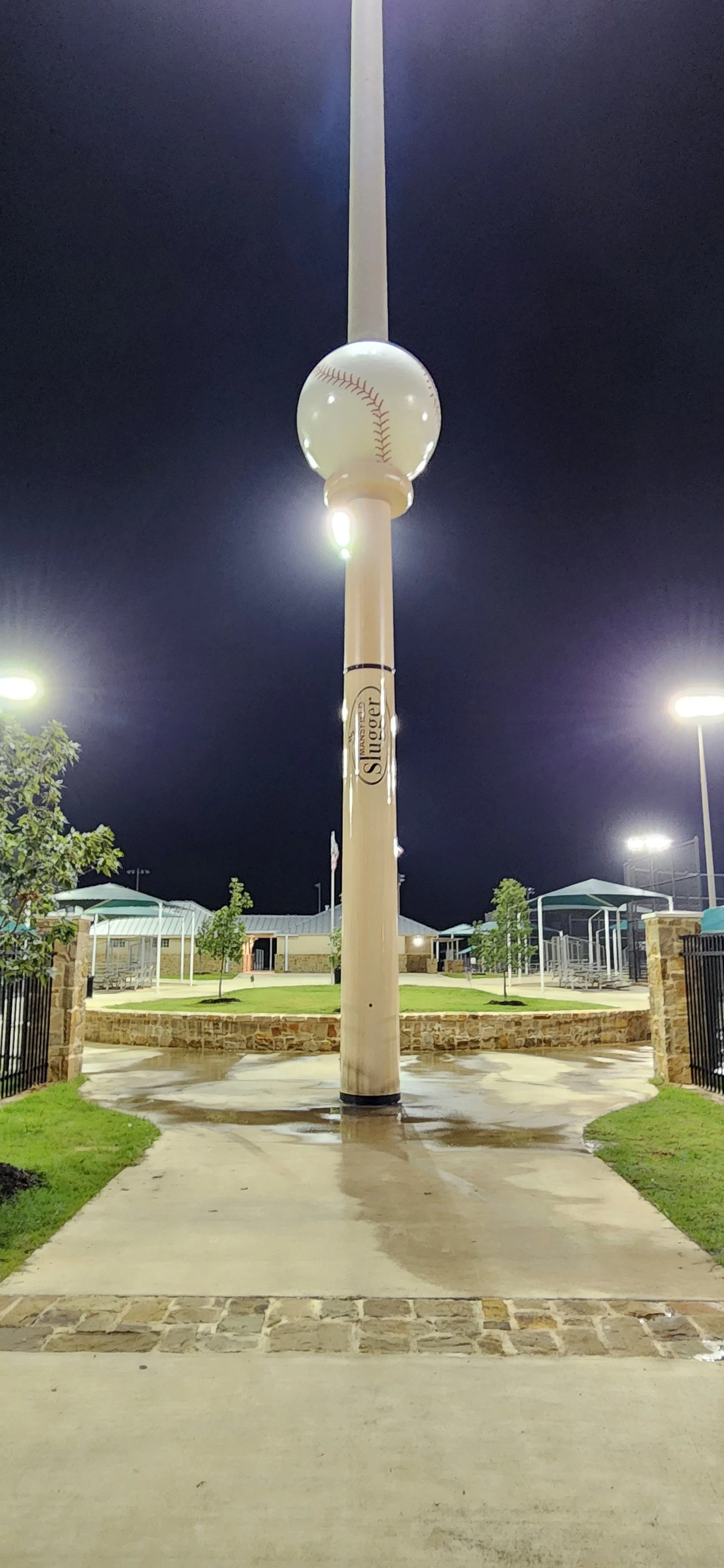 A large pole with a baseball-shaped decoration at the top, resembling a home run indicator, in a park at night. Surrounding the pole are pathways, trees, and park amenities.