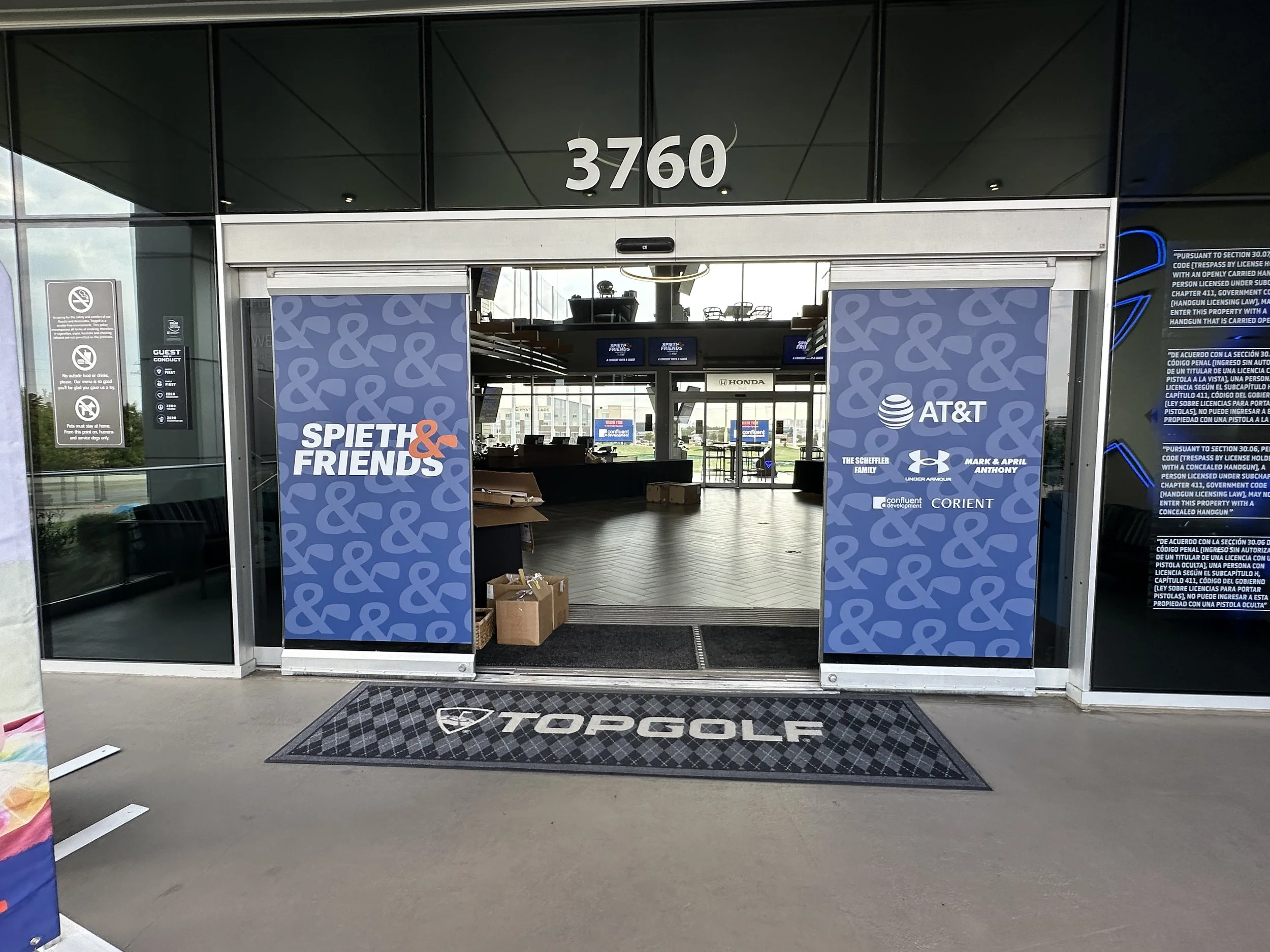 Entrance to a golf facility with blue banners displaying the words 'SPIETH & FRIENDS' and sponsor logos, including AT&T, under Armour, and others. The address number 3760 is above the entrance, and a black doormat with 'TOPGOLF' logo is on the ground.