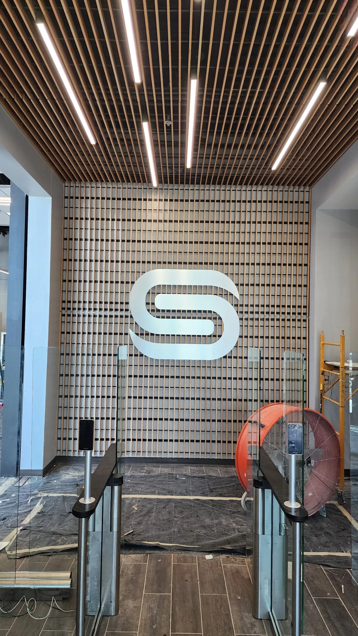 Interior of a modern building with wood paneling, ceiling lights, a white logo on the wall, and automated entrance gates.