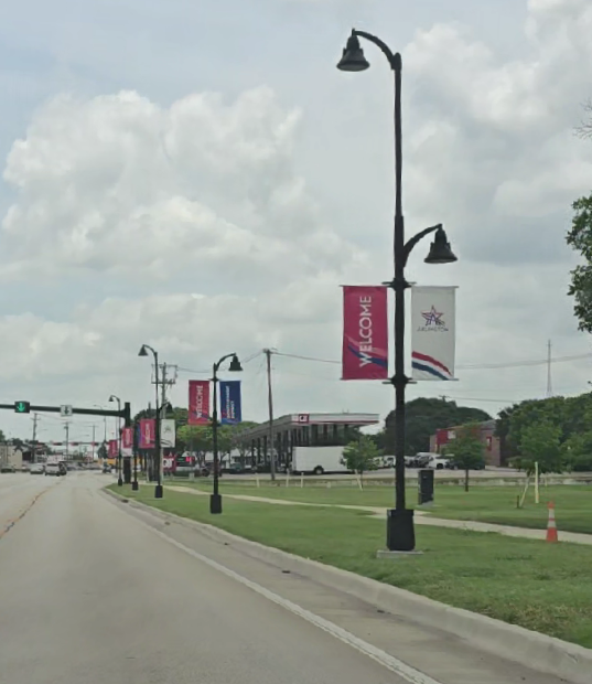 Street view with lamp posts and banners welcoming visitors, with a gas station and some buildings in the background under cloudy sky.