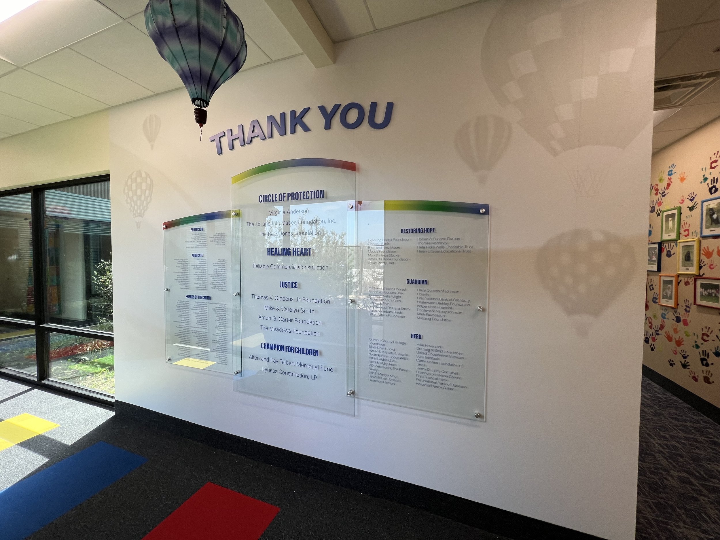 Wall display with the words 'Thank You' in large letters, decorative hanging hot air balloons, and plaques with lists of names and titles in a hallway with colorful handprints on the adjacent wall.