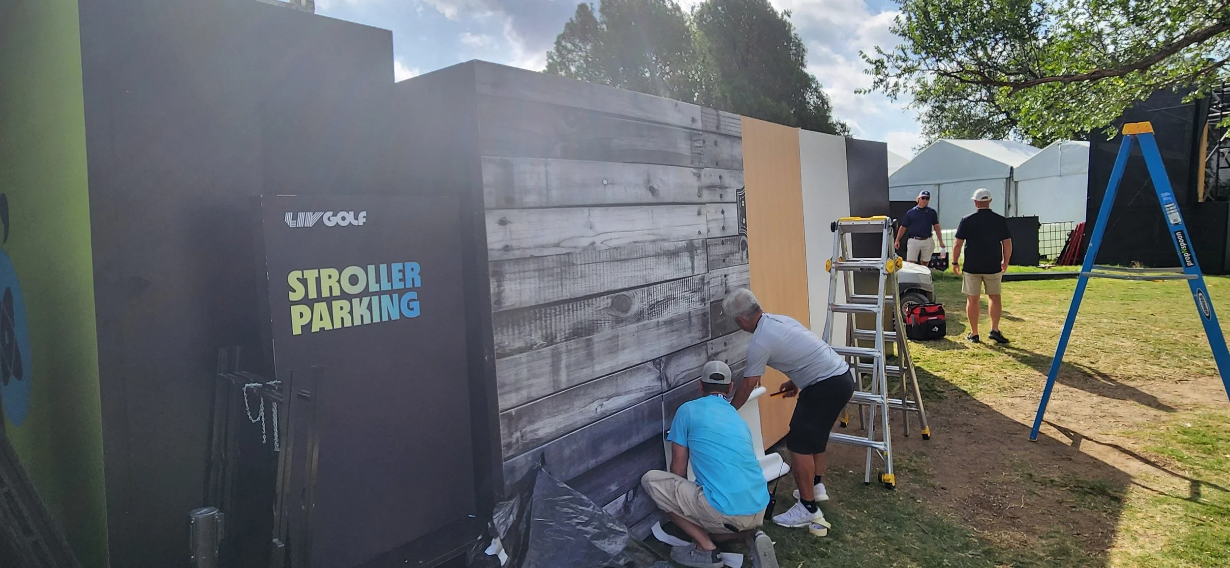 People setting up a display booth with a wooden and gray panel at an outdoor event, with a ladder and various equipment around, under a partly cloudy sky with trees in the background.