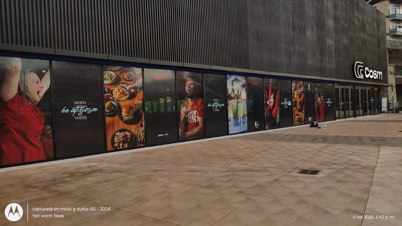 Exterior view of a commercial building with large windows displaying advertisements, including images of food, people celebrating, and beverages, with the word 'Cosm' on the facade.