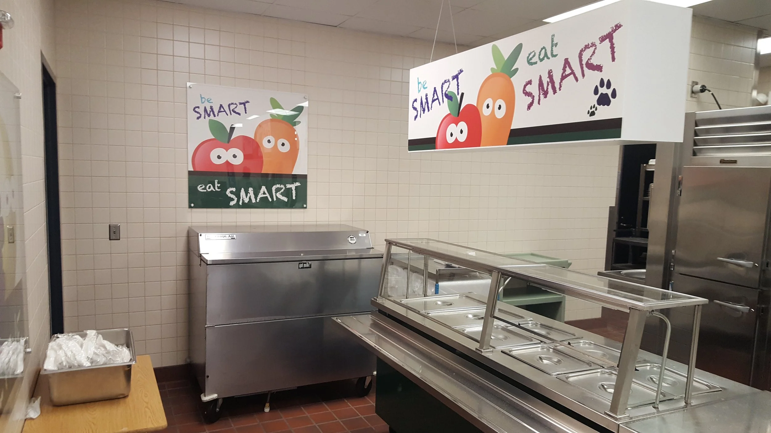 Empty cafeteria serving area with metal food trays, signs on the wall encouraging healthy eating, and a tiled wall in the background.