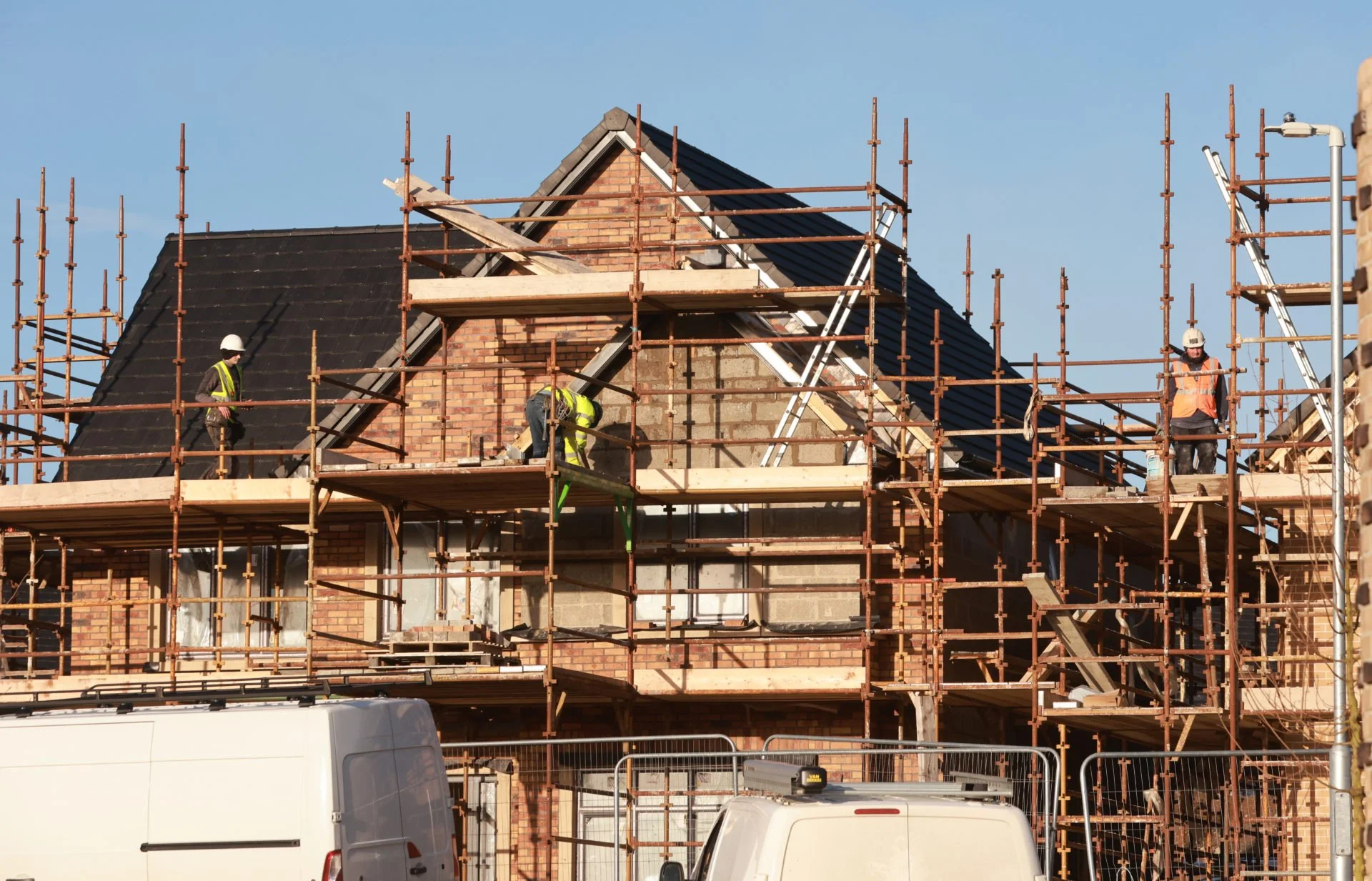 Construction workers on scaffolding working on a brick house roof under clear blue sky.