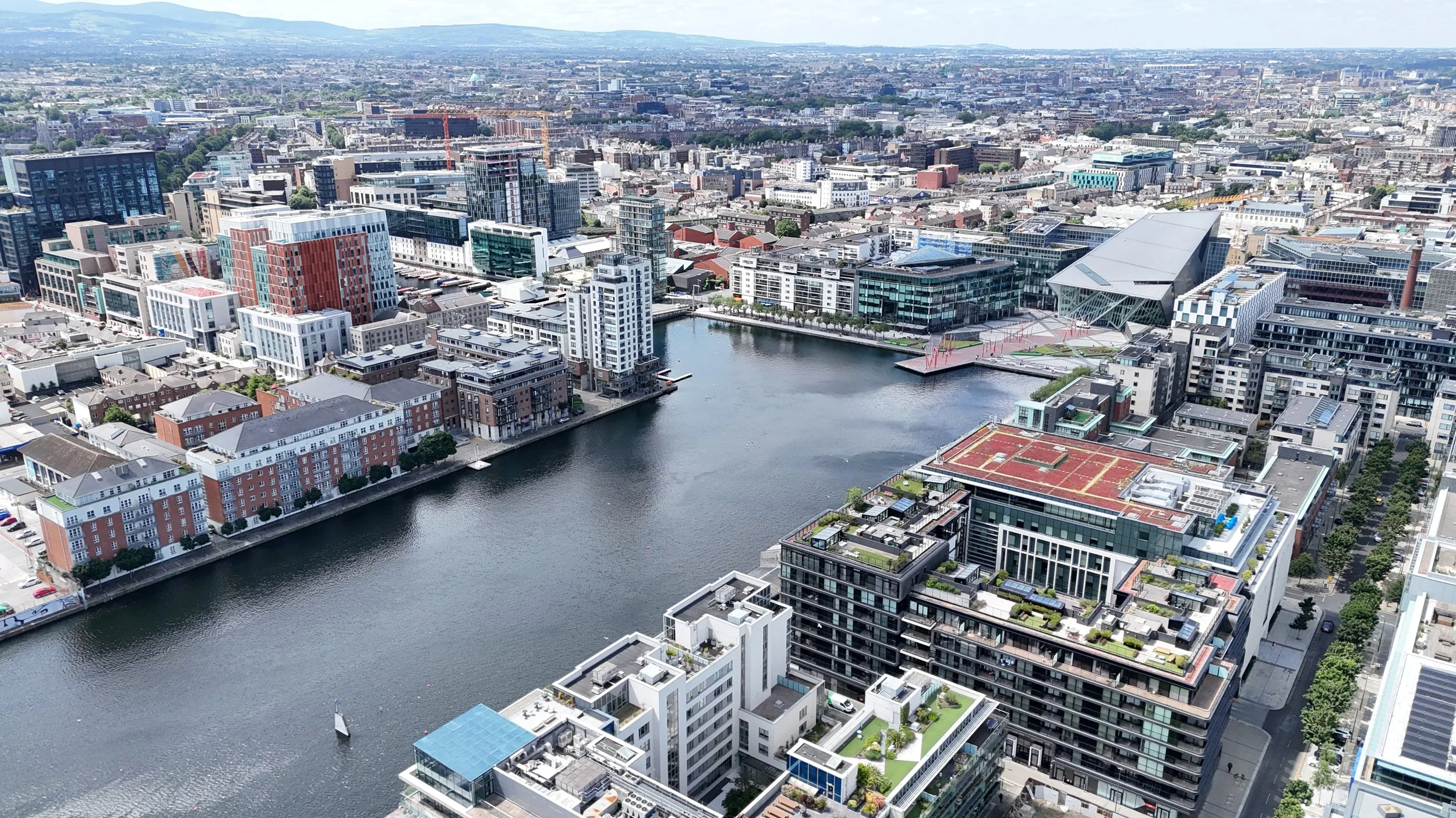 Aerial view of a cityscape featuring modern high-rise buildings, a waterway with boats, and a skyline with mountains in the background.
