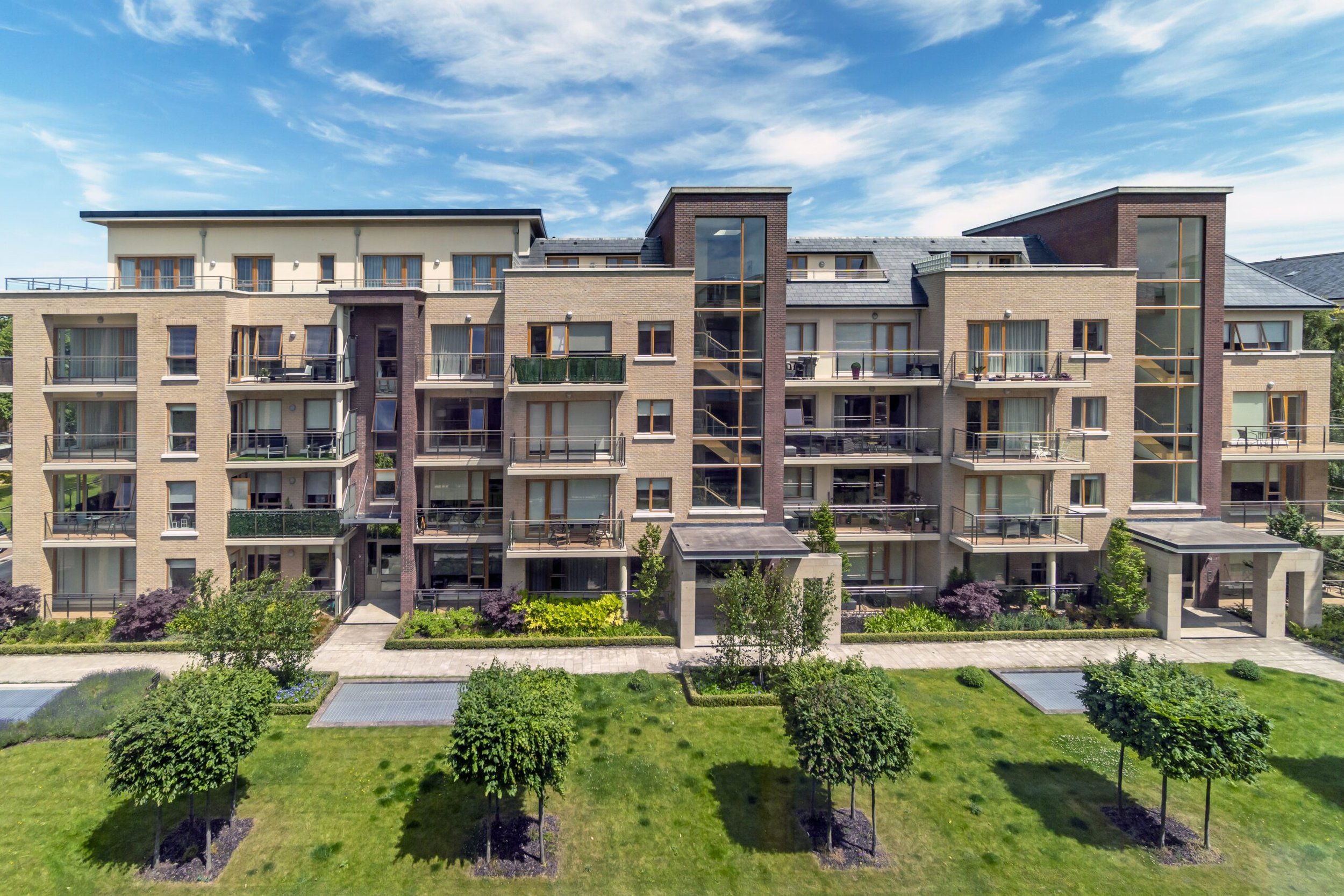A modern multi-story residential apartment building with balconies, large windows, and a landscaped garden in the foreground under a partly cloudy sky.