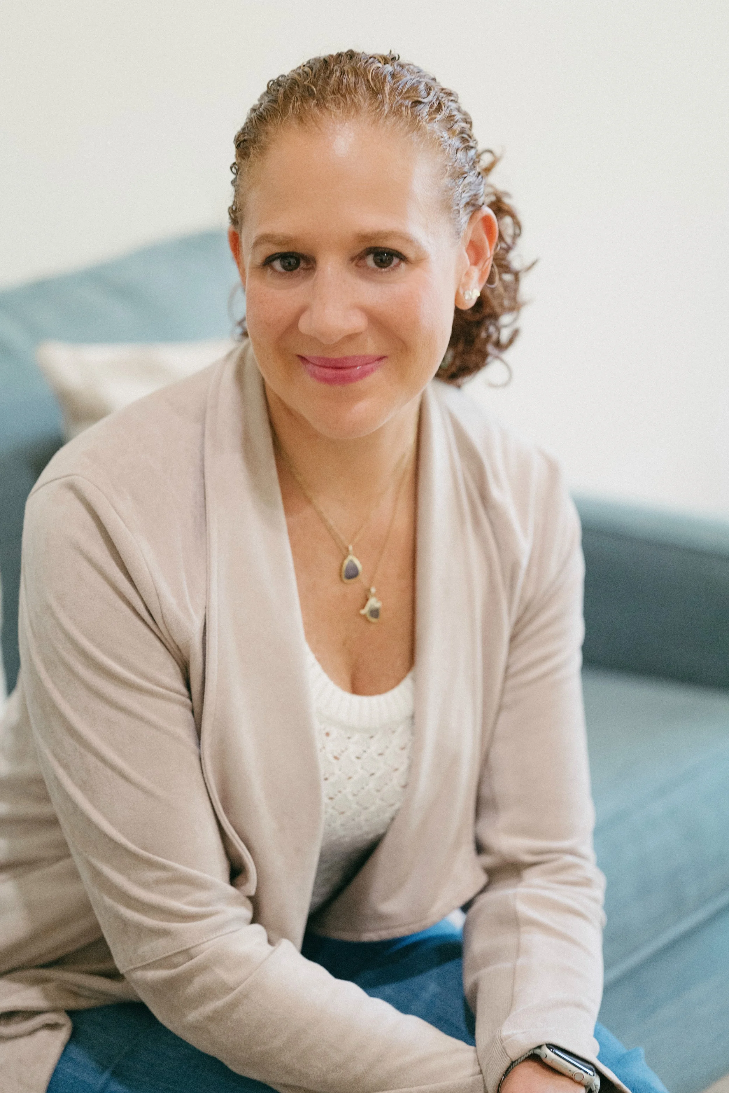 A woman with curly red hair and light skin smiling while sitting on a blue couch, wearing a beige blazer, white top, and layered necklaces.
