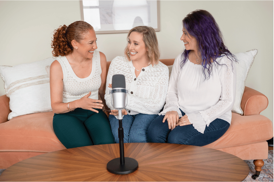 Three women sitting on a peach-colored sofa, smiling and talking, with a microphone on a wooden table in front of them, in a bright living room.