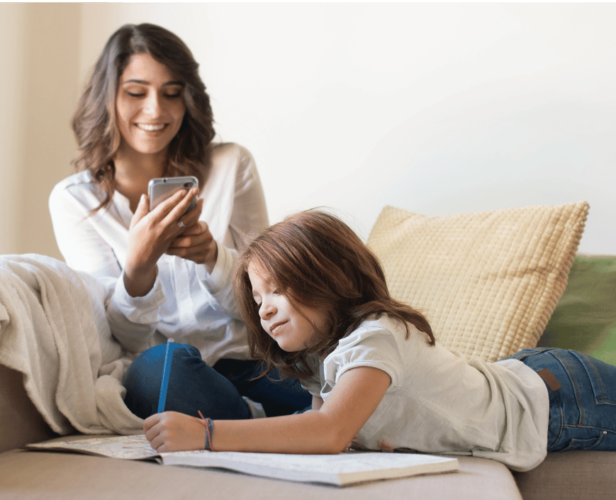 Young woman sitting on a couch taking a photo with her phone while a girl lies on her stomach drawing or writing on a large notebook on her lap.