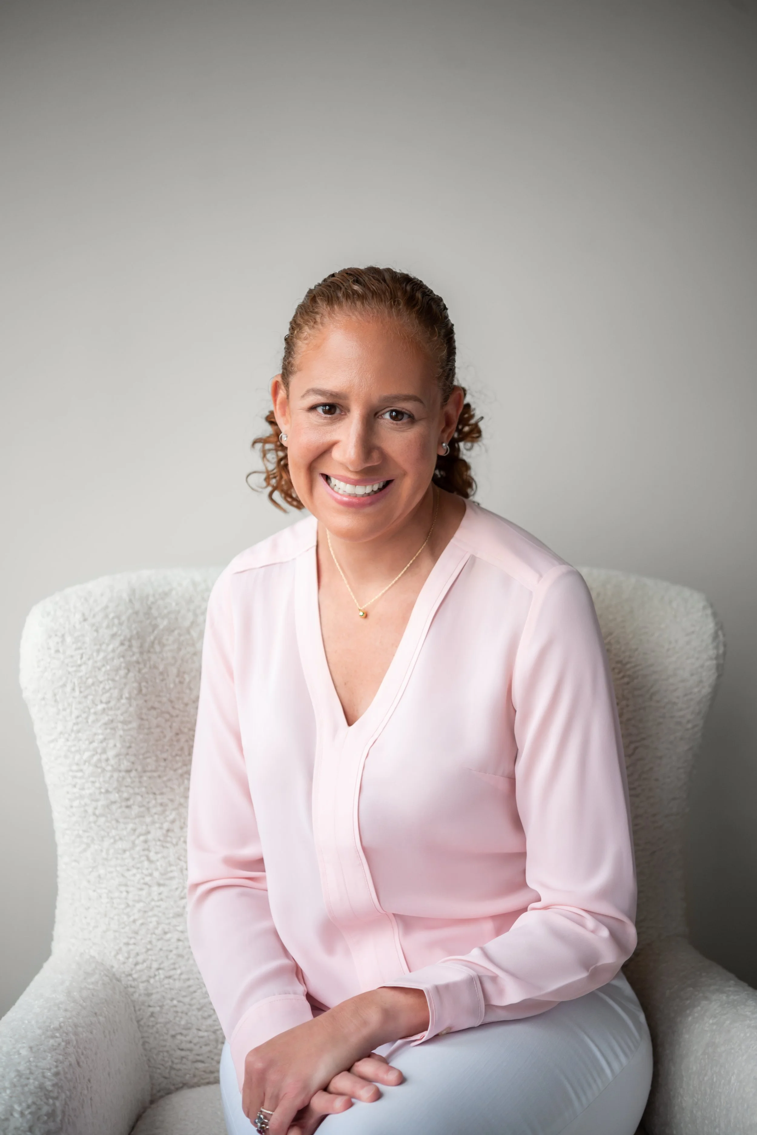 Portrait of a woman with curly hair smiling, sitting on light-colored armchair, wearing a light pink blouse and jewelry, against a plain gray background.