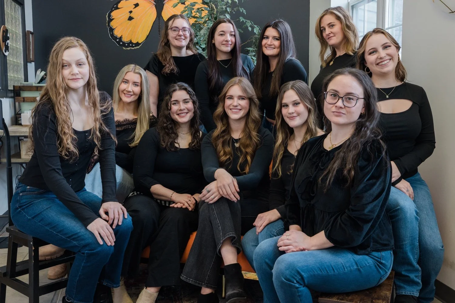 A group of eleven women gathered in an indoor space with a butterfly mural on the wall behind them, smiling at the camera.