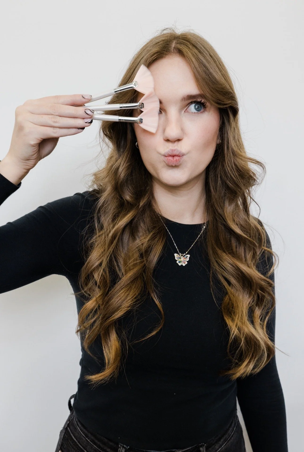 A woman with long, wavy brown hair is holding makeup brushes in front of her face, with some brushes on her forehead, and making a playful expression.
