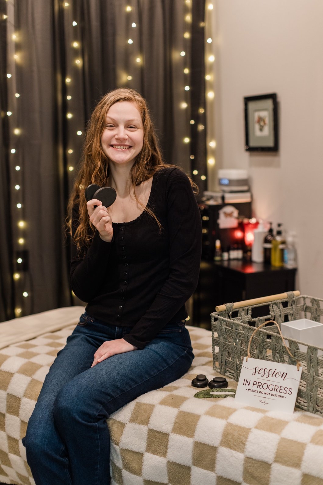 Woman smiling and sitting on a massage table in a decorated room holding two black stones. There is a sign on the table that reads 'session in progress'.
