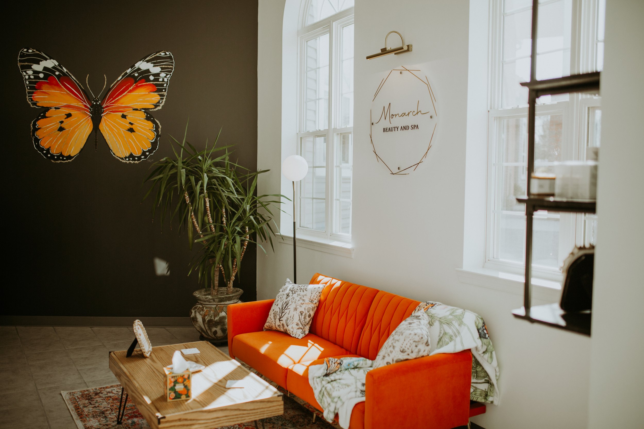 Modern interior of a beauty and spa salon with an orange sofa, decorative pillows, a wooden coffee table, a potted plant, and butterfly wall art.