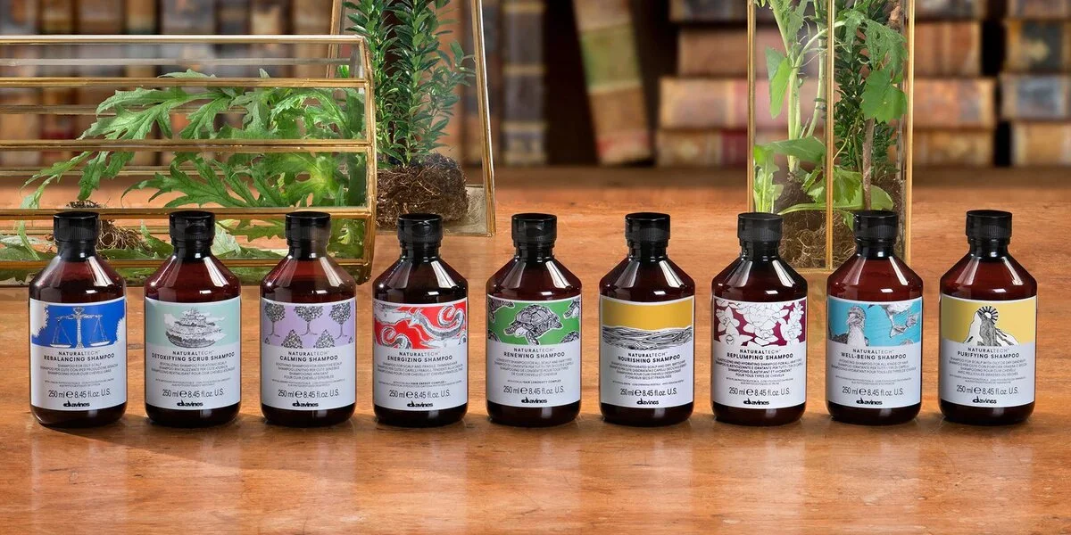 A row of eight brown bottles of Davines shampoo on a wooden surface, each with a colorful label and black cap, with plants and a wooden bookshelf in the background.