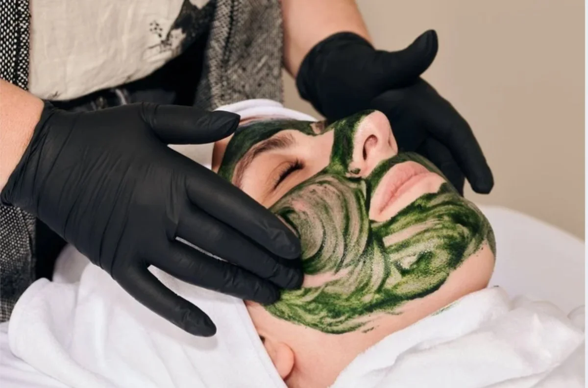 Person lying down with a green patterned face mask being applied by a person wearing black gloves.