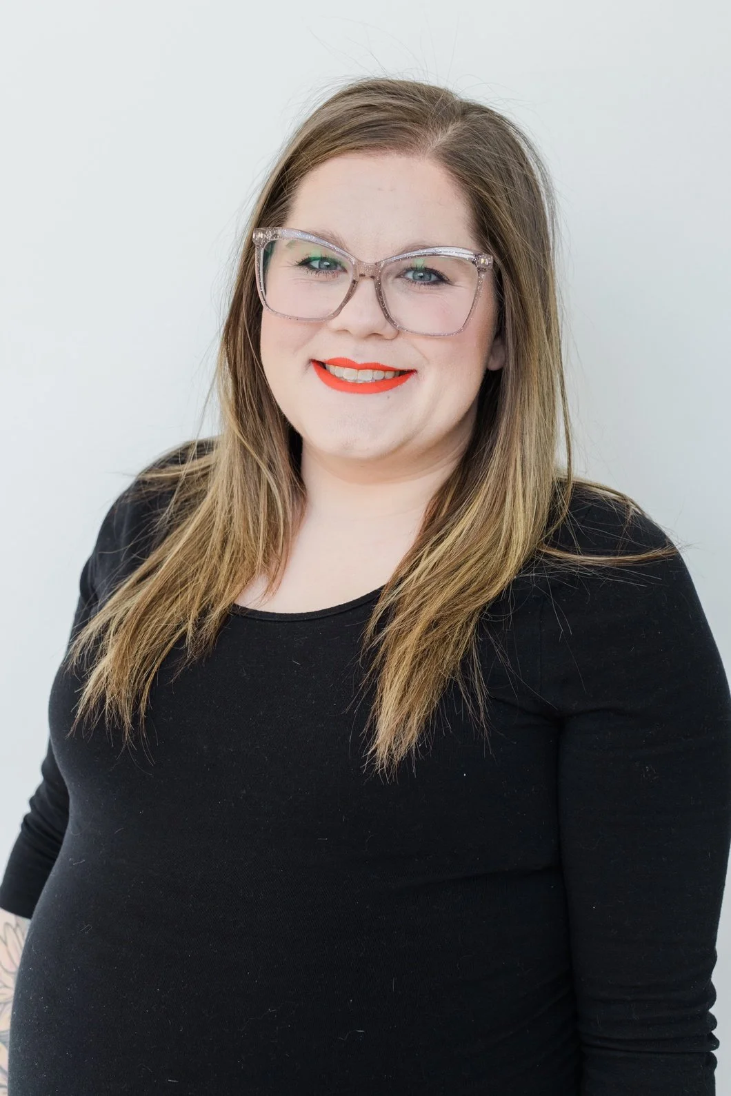 A woman with long brown hair, wearing transparent glasses, red lipstick, and a black top, smiling at the camera, against a plain white background.