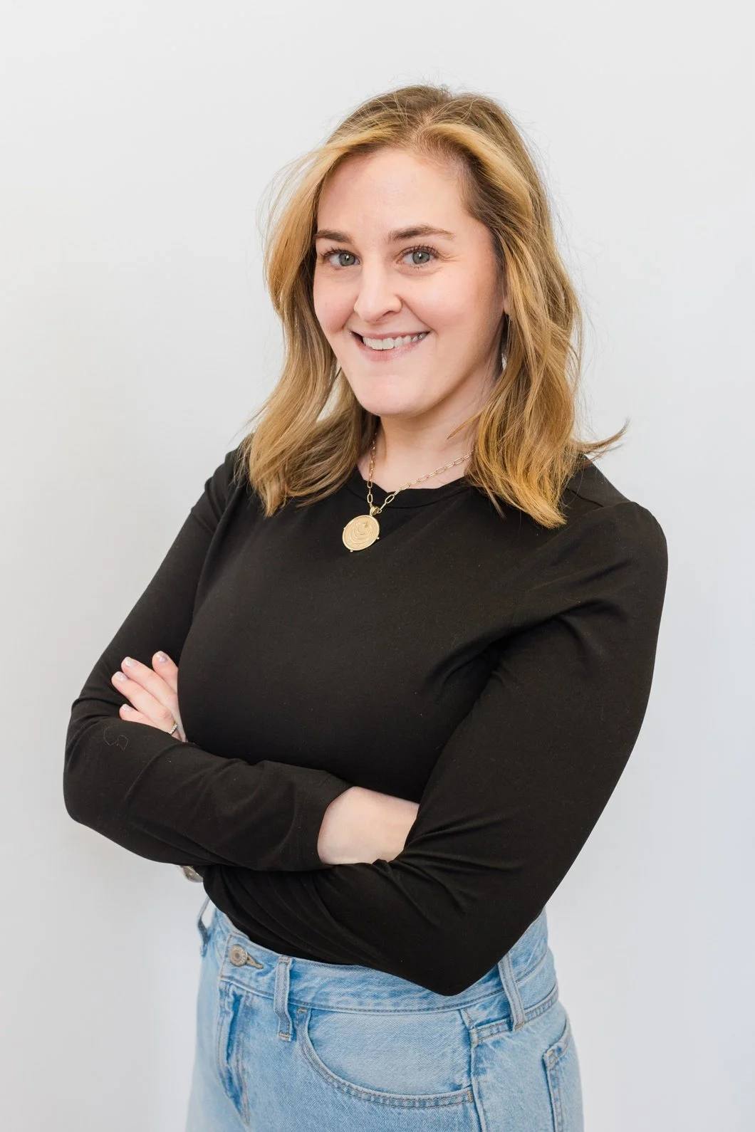 A woman with shoulder-length blonde hair, wearing a black long-sleeve shirt, light blue jeans, and a gold necklace, smiling with her arms crossed against a plain white background.