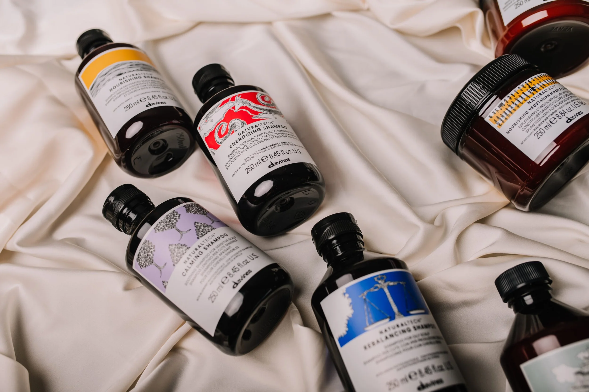 Several bottles of hair care products, including shampoo and conditioner, are scattered on a cream-colored fabric surface.