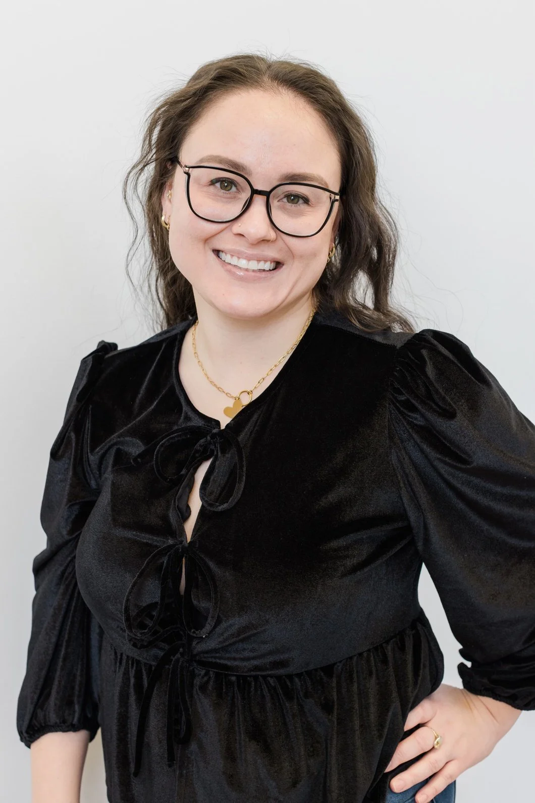 A young woman smiling, wearing glasses, a black velvet top with bows, and gold jewelry against a plain light-colored background.
