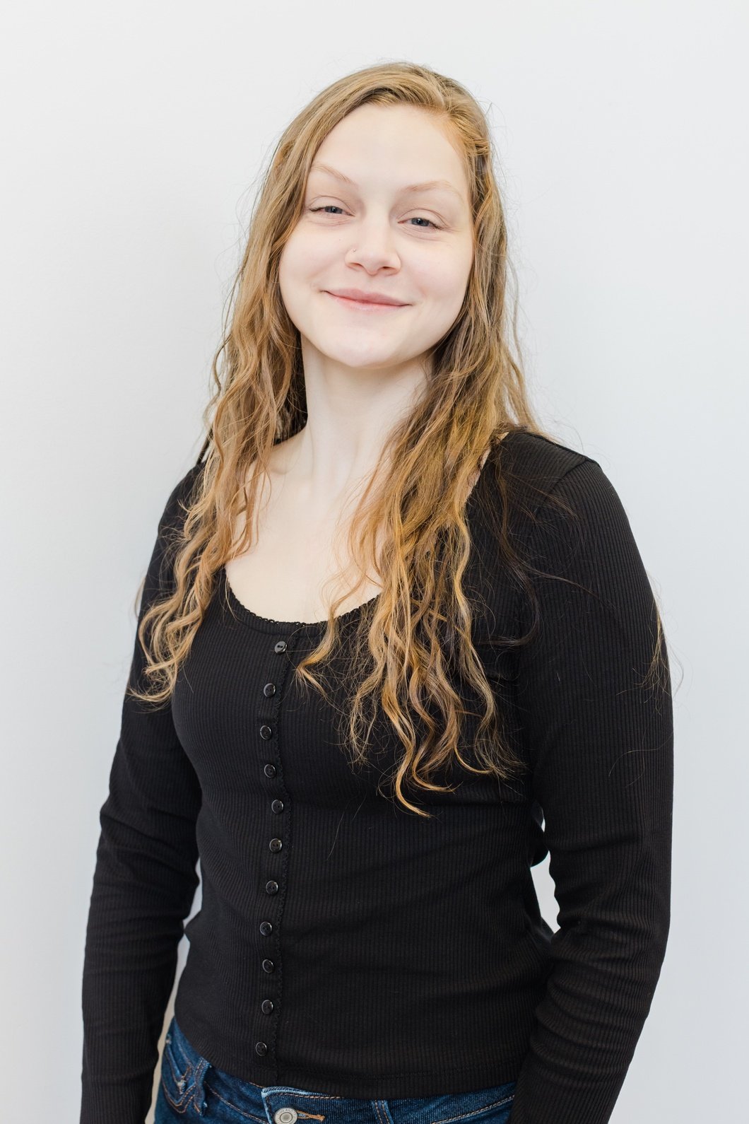 A young woman with long, wavy red hair smiling and looking at the camera, wearing a black long-sleeve button-up top and blue jeans, standing against a plain white background.