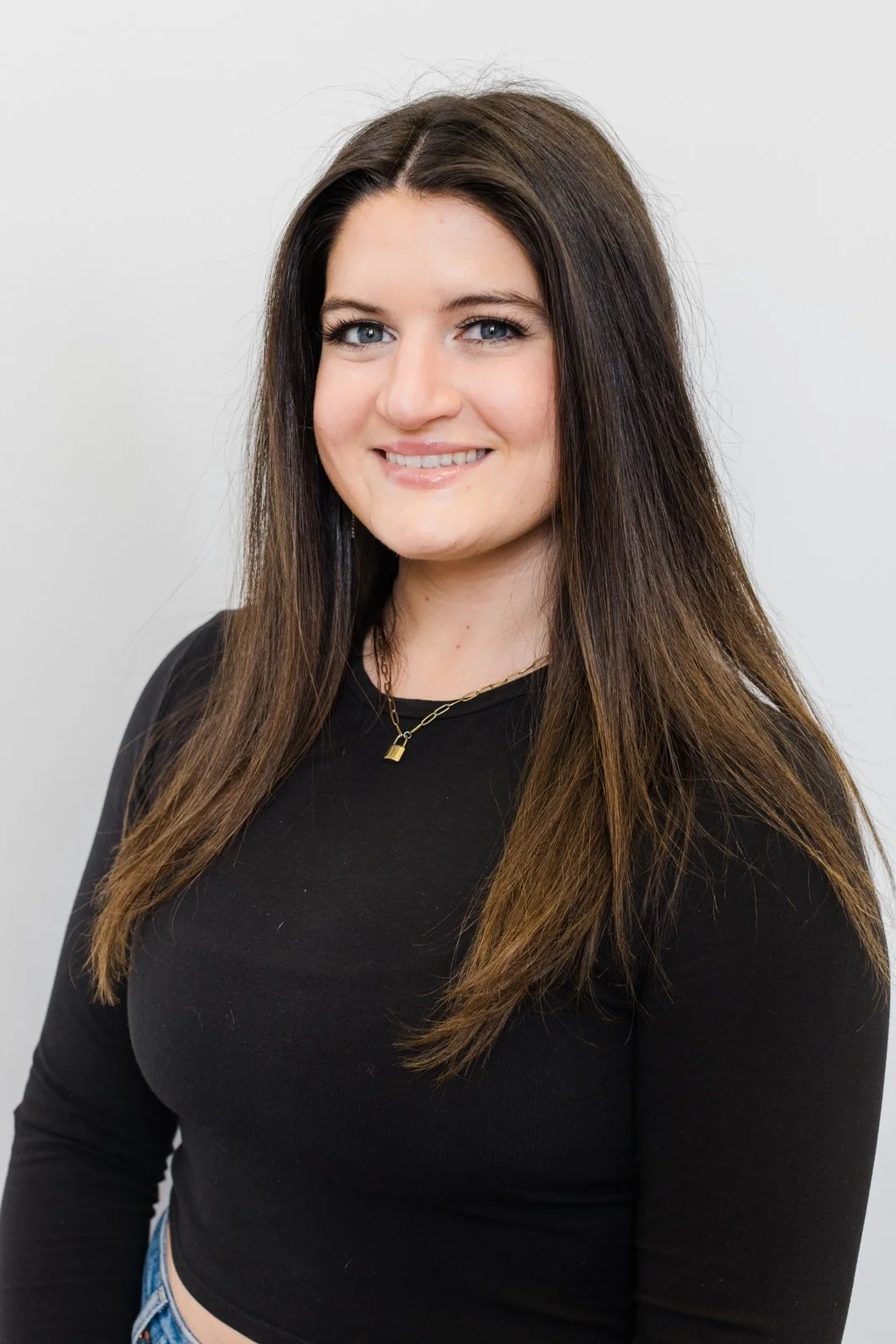 A young woman with long brown hair, blue eyes, and a smile, wearing a black long-sleeve shirt and a gold necklace with a lock pendant, standing against a plain white background.