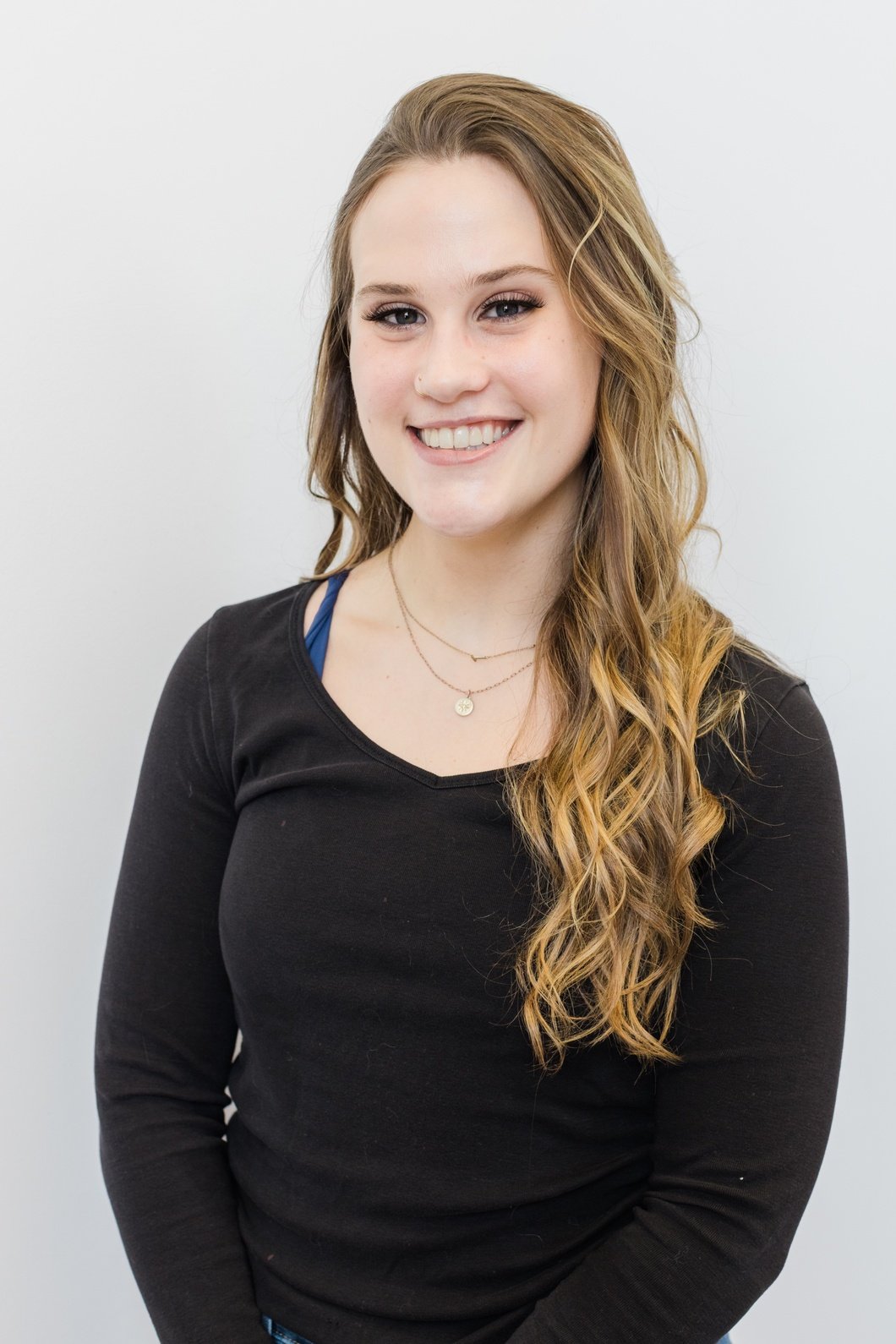 A young woman with long wavy light brown hair, smiling, wearing a black long-sleeve shirt and layered necklaces, standing against a plain light gray background.