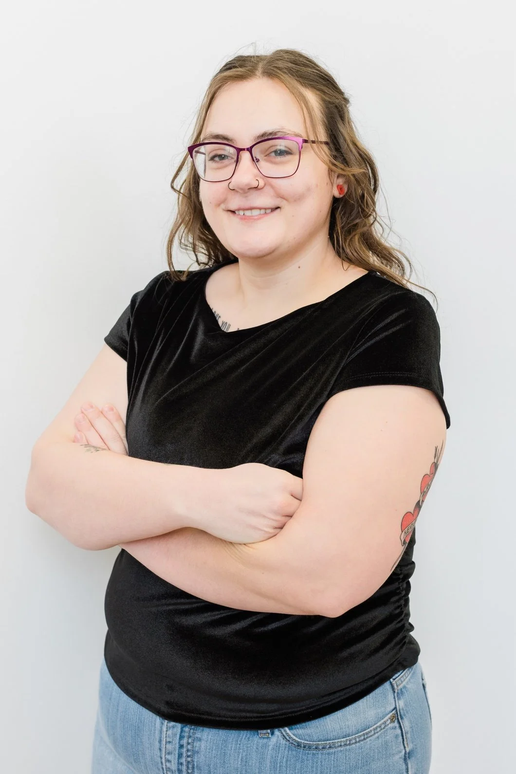 A young woman with glasses, curly hair, and tattoos, wearing a black velvet top and jeans, standing with arms crossed and smiling in front of a plain white background.