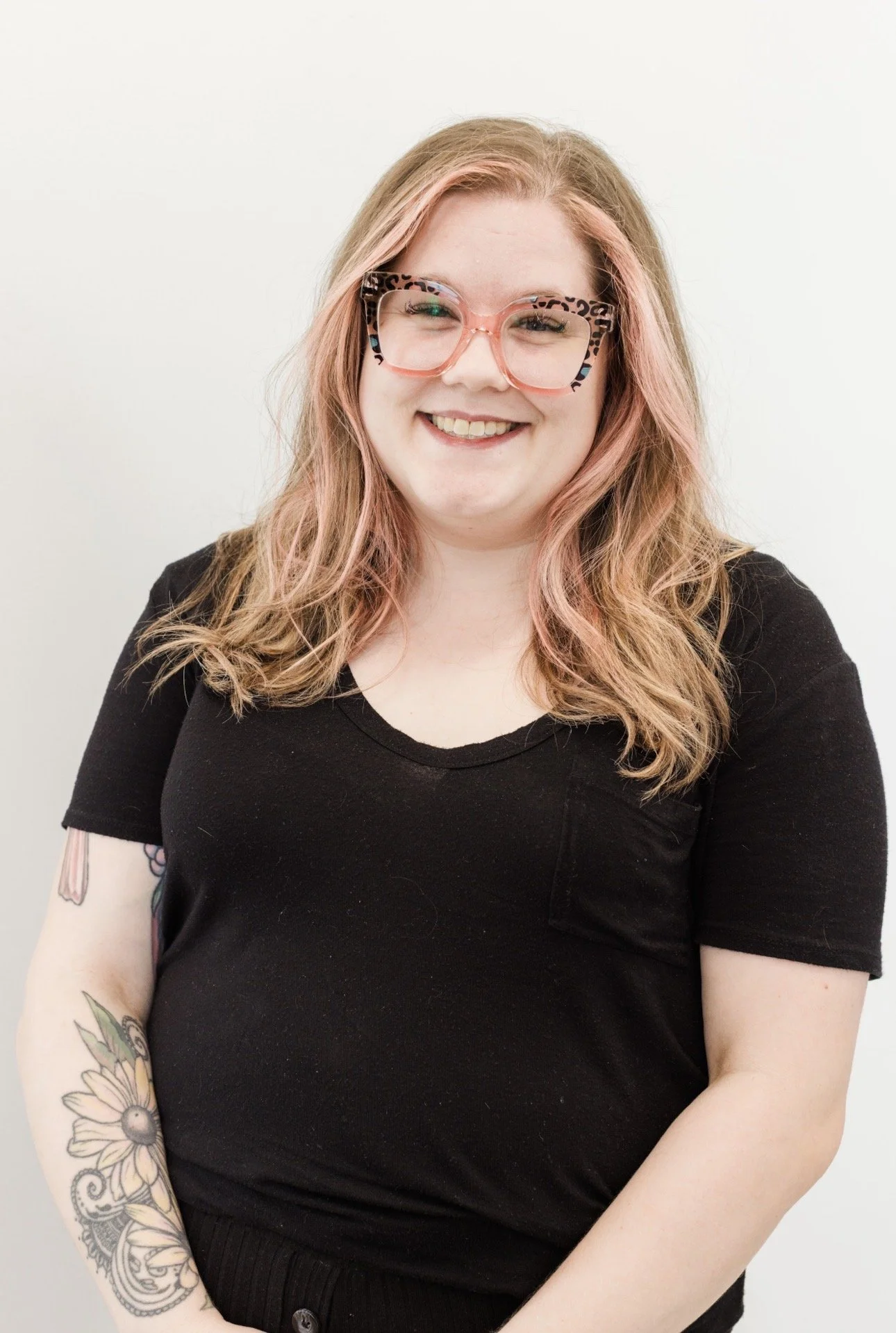 A woman with long brown hair, wearing transparent glasses, red lipstick, and a black top, smiling at the camera, against a plain white background.