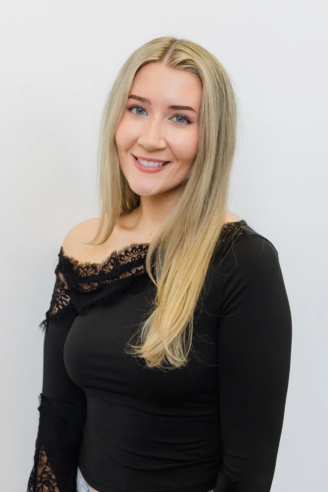 A woman with long blonde hair, blue eyes, and fair skin, smiling, wearing a black lace-shoulder top, poses against a plain white background.