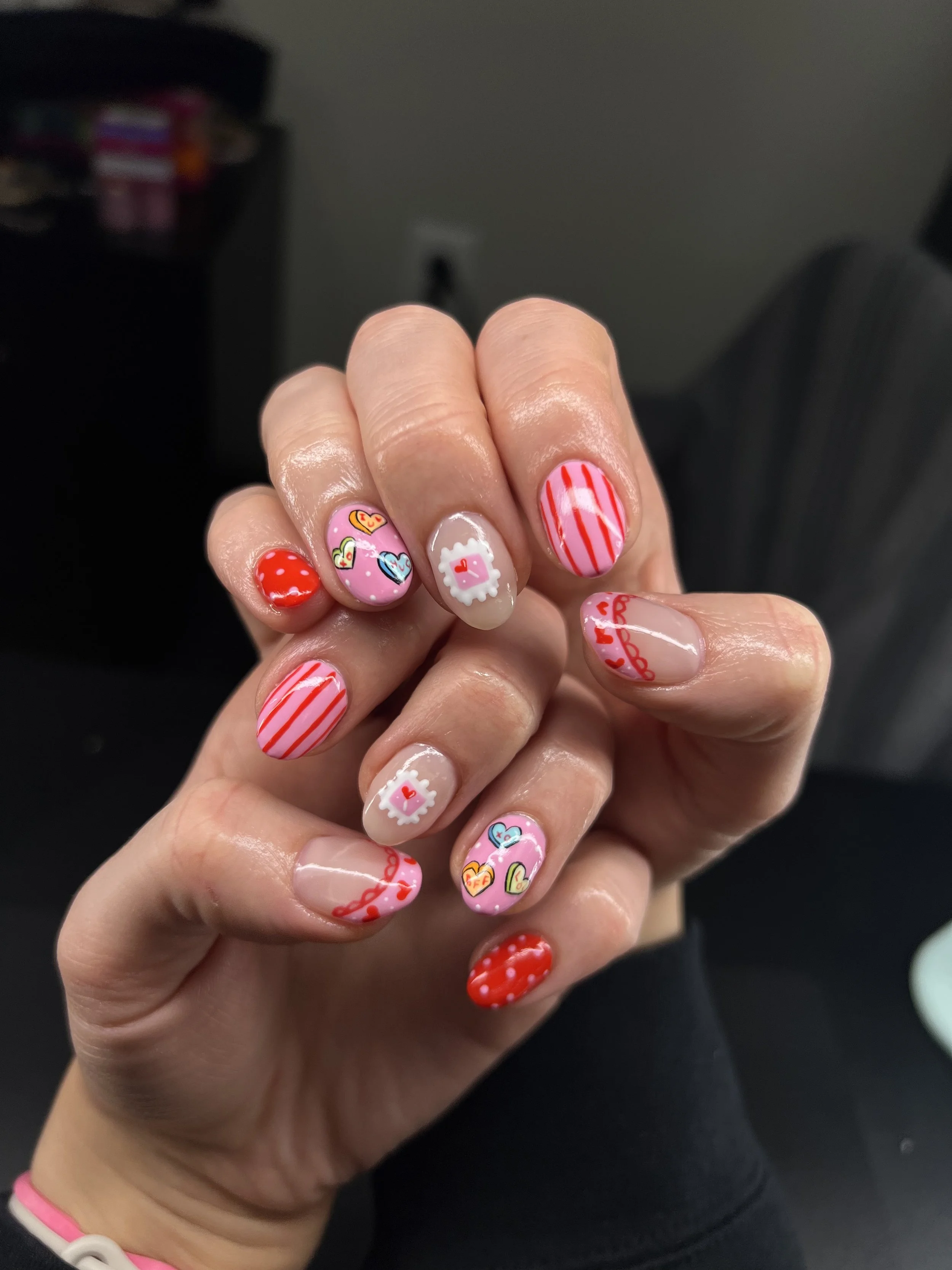Hand with decorated Valentine's Day themed nail art, featuring hearts, stripes, and love-related designs in red, pink, and white colors.