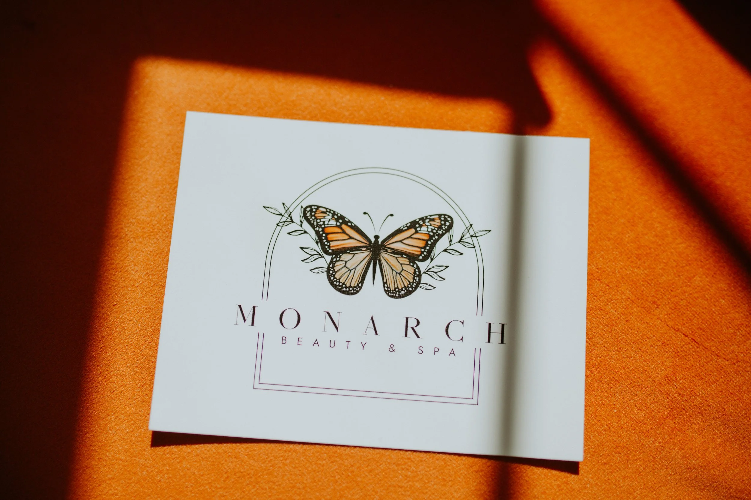 A white card with a butterfly illustration and the words "Monarch Beauty & Spa" on an orange surface with shadows.