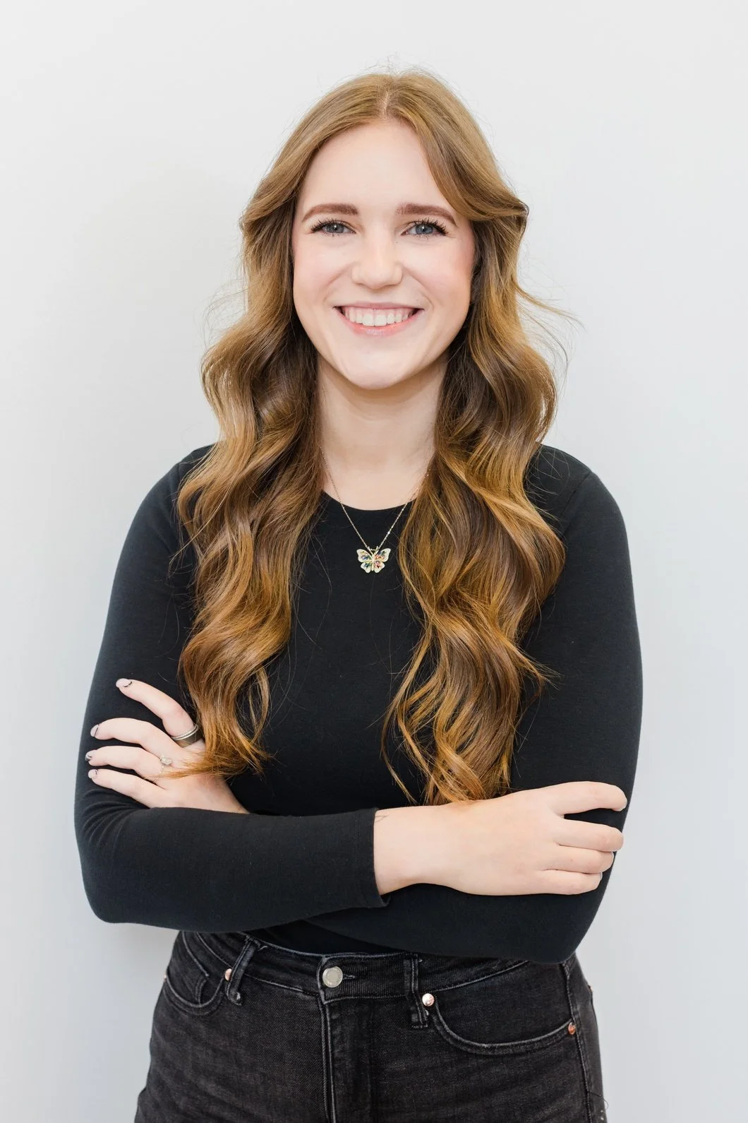 Young woman with long, wavy red hair wearing a black long-sleeve top, crossing her arms, smiling, standing against a plain light gray background.