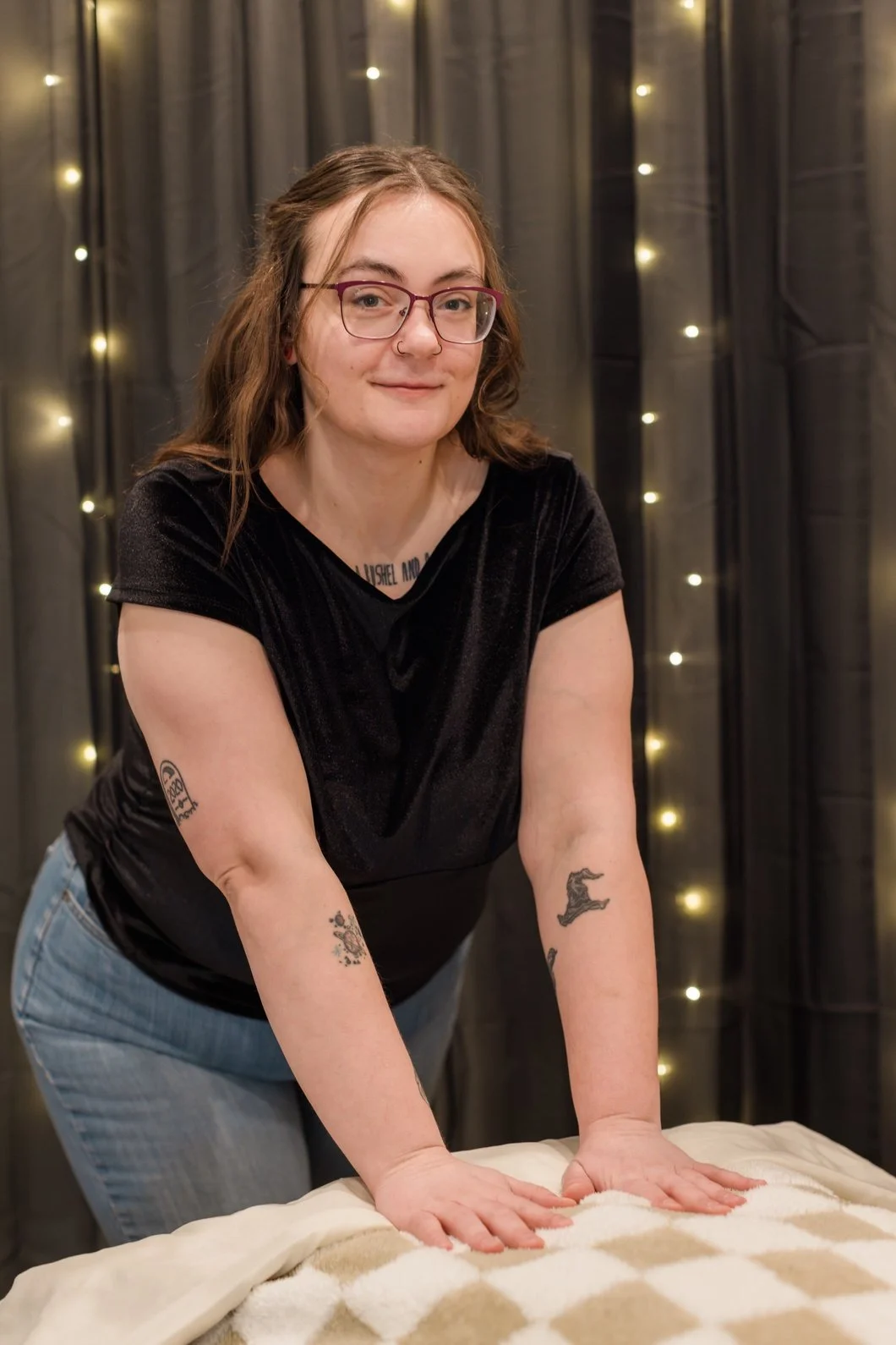 A young woman with glasses, tattoos, and a nose piercing leaning forward on a bed with string lights and dark curtains in the background.